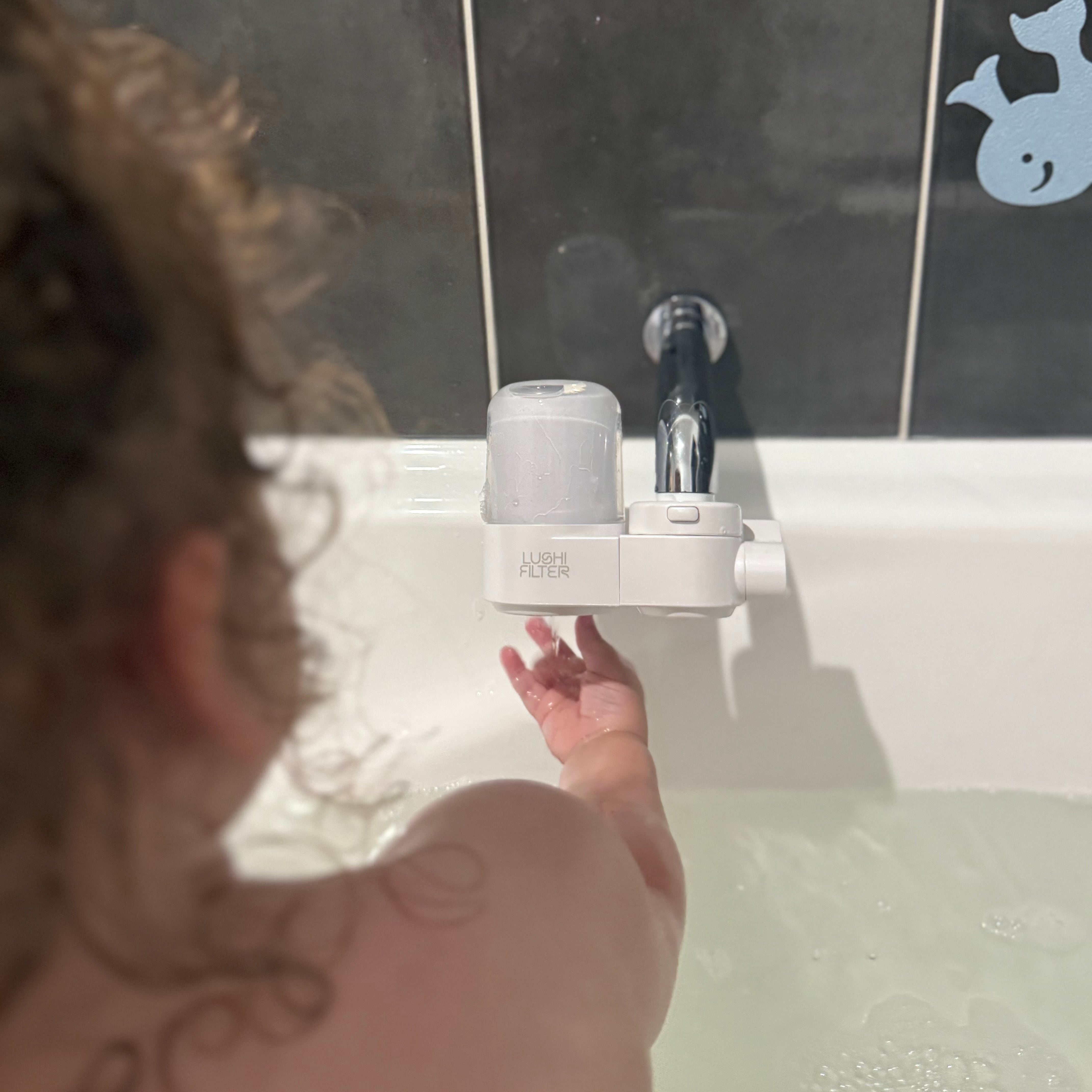 Bath tub filter kid feeling the filtered water, white water filter device against a tiled bathroom wall.